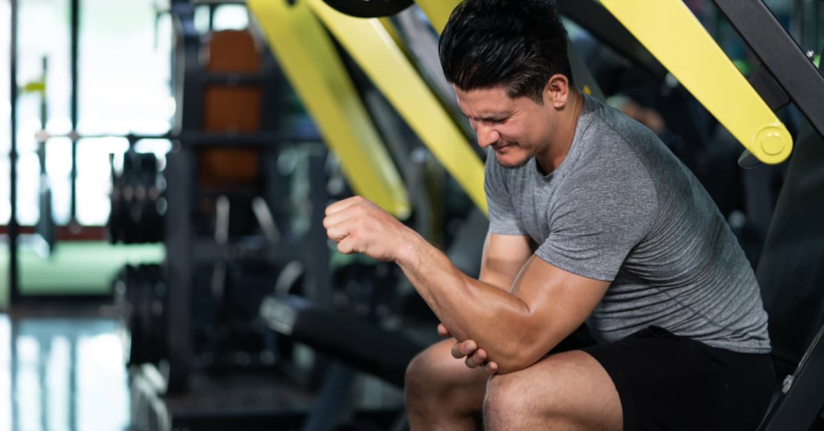 man wearing a gray shirt in the gym holding his elbow while wincing in pain in the gym